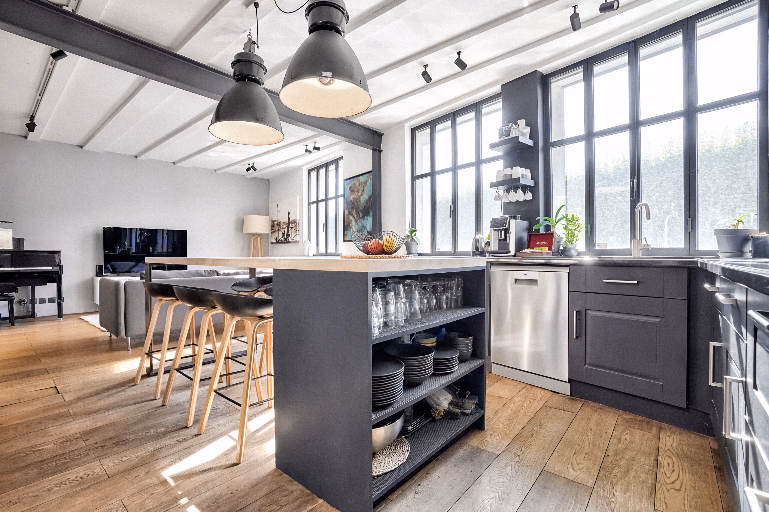 Cuisine équipée du loft industriel avec îlot central, verrières type atelier, luminaires suspendus et espace salon ouvert pour productions et événements