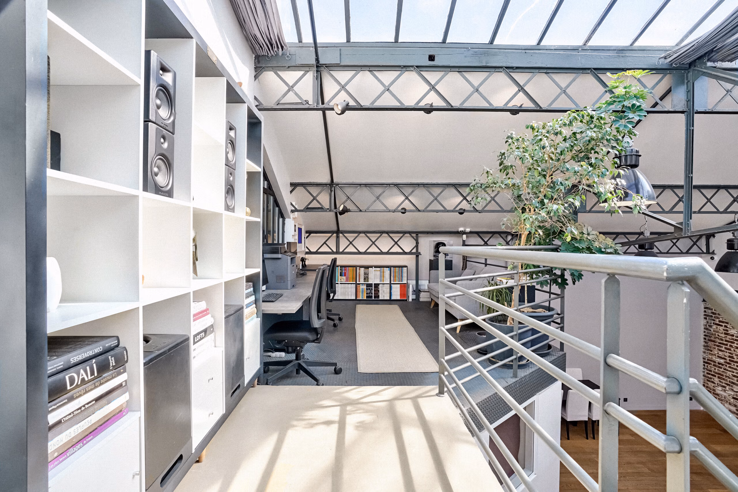Mezzanine bureau du loft avec poutres métalliques apparentes, verrière industrielle, bibliothèque et poste de travail sous lumière naturelle pour réunions et tournages