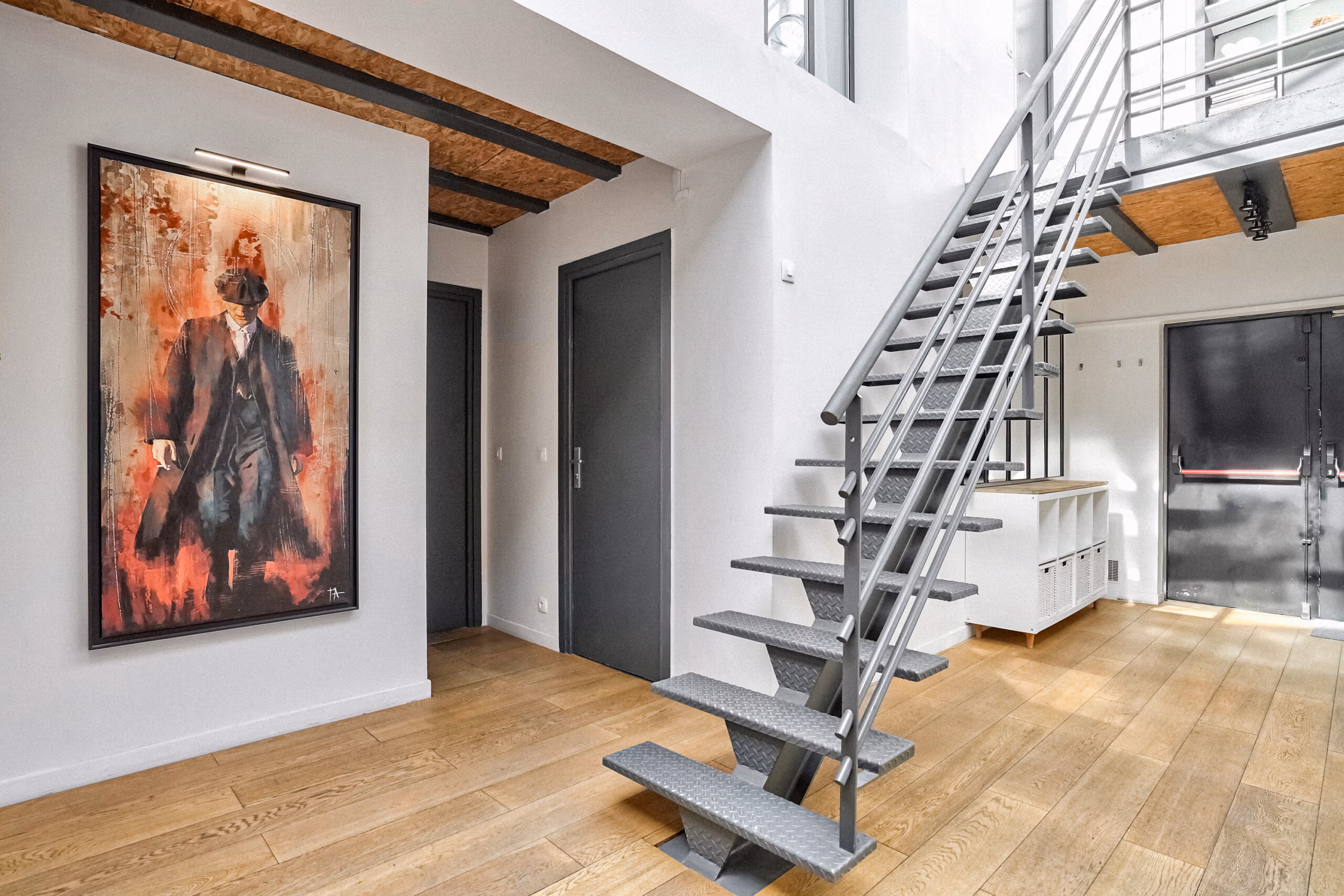Escalier métallique design avec œuvres d'art encadrées, poutres apparentes et parquet bois, reflétant l'identité artistique du lieu pour shootings créatifs et tournages d'ambiance galerie