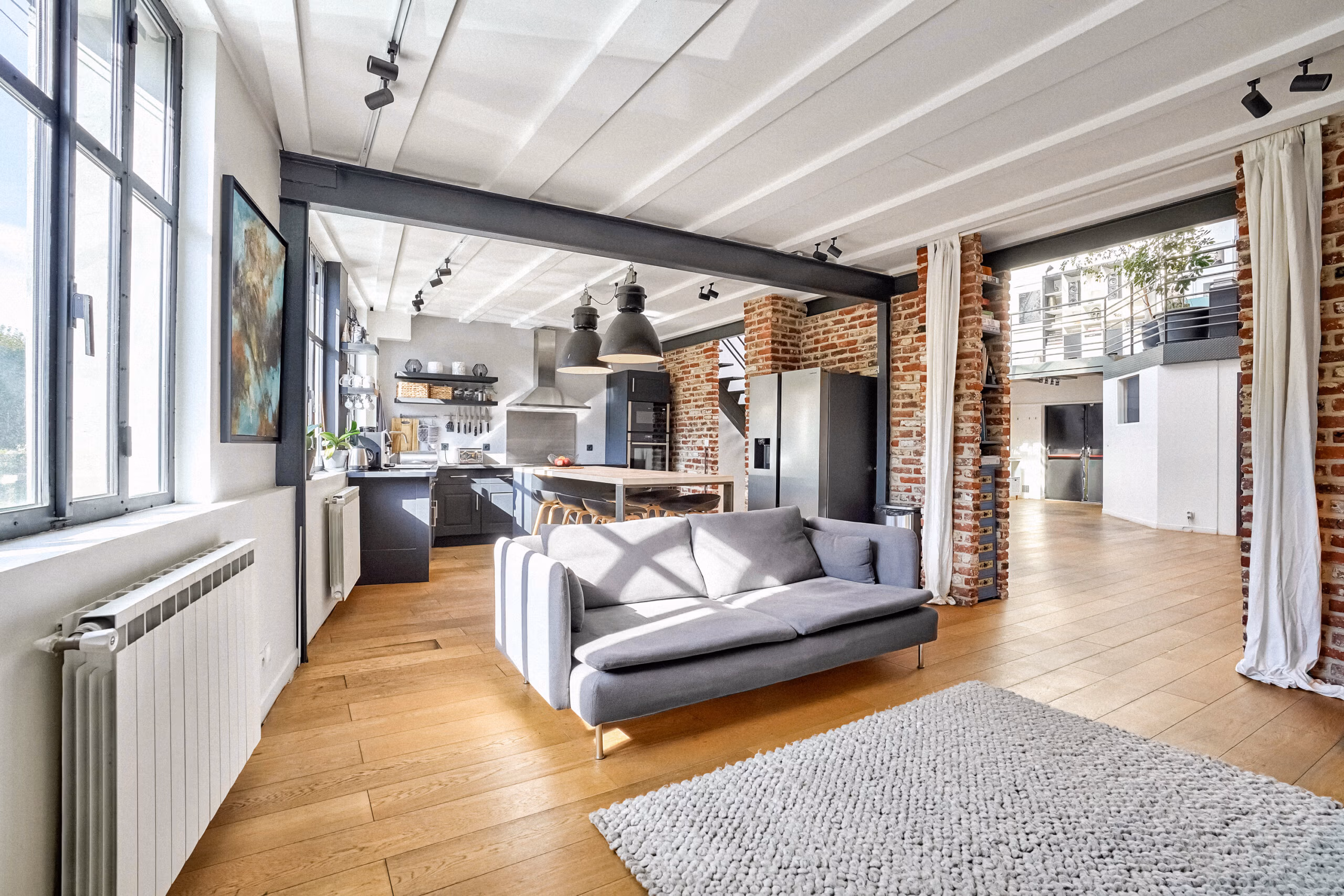 Open space avec cuisine équipée ouverte sur salon, colonnes de briques structurantes, poutres peintes blanches et lumière naturelle pour tournages publicitaires et contenus lifestyle