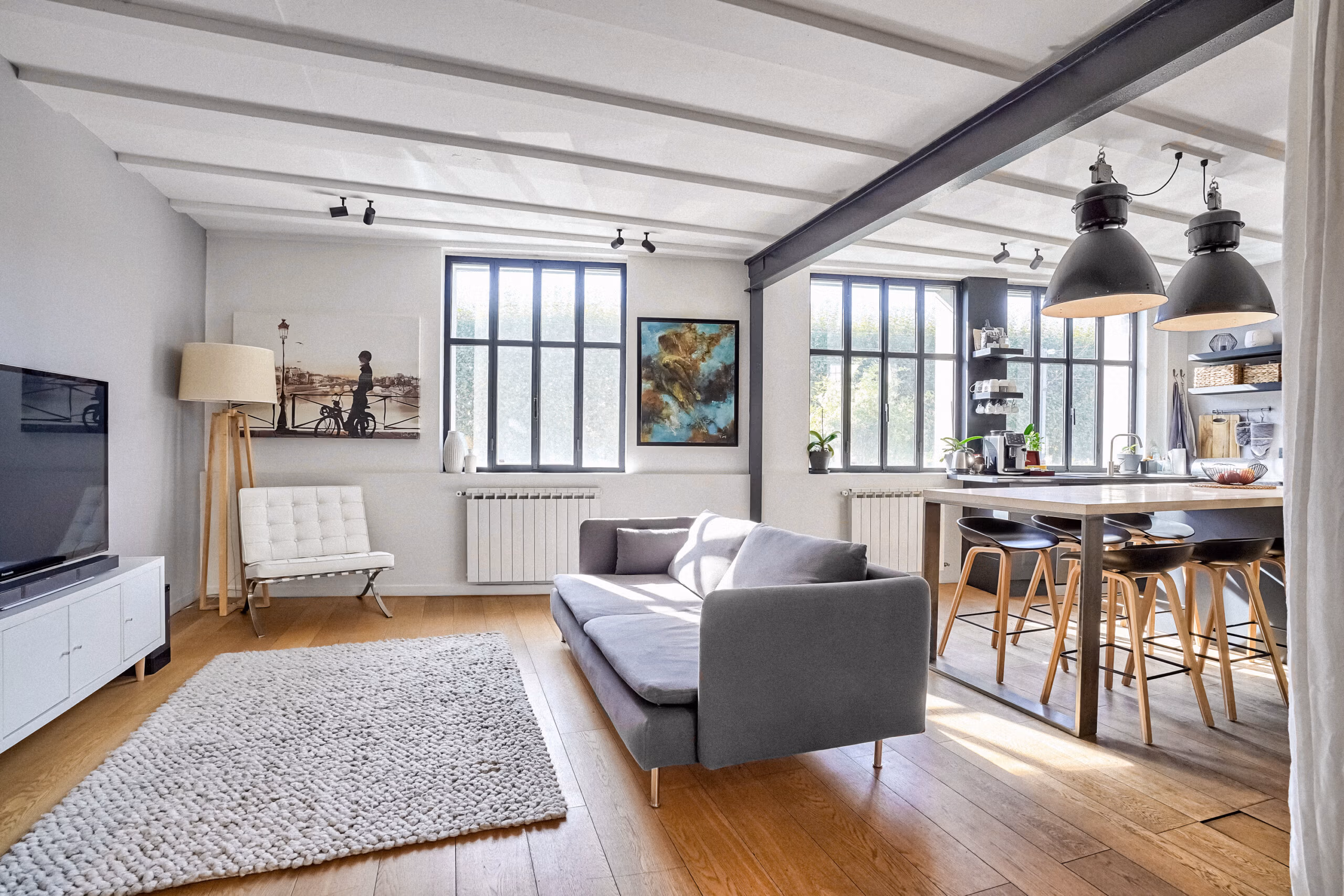 Vue panoramique du salon-cuisine avec verrières type atelier, luminaires industriels XXL, mobilier design et œuvres murales pour shootings déco et films institutionnels