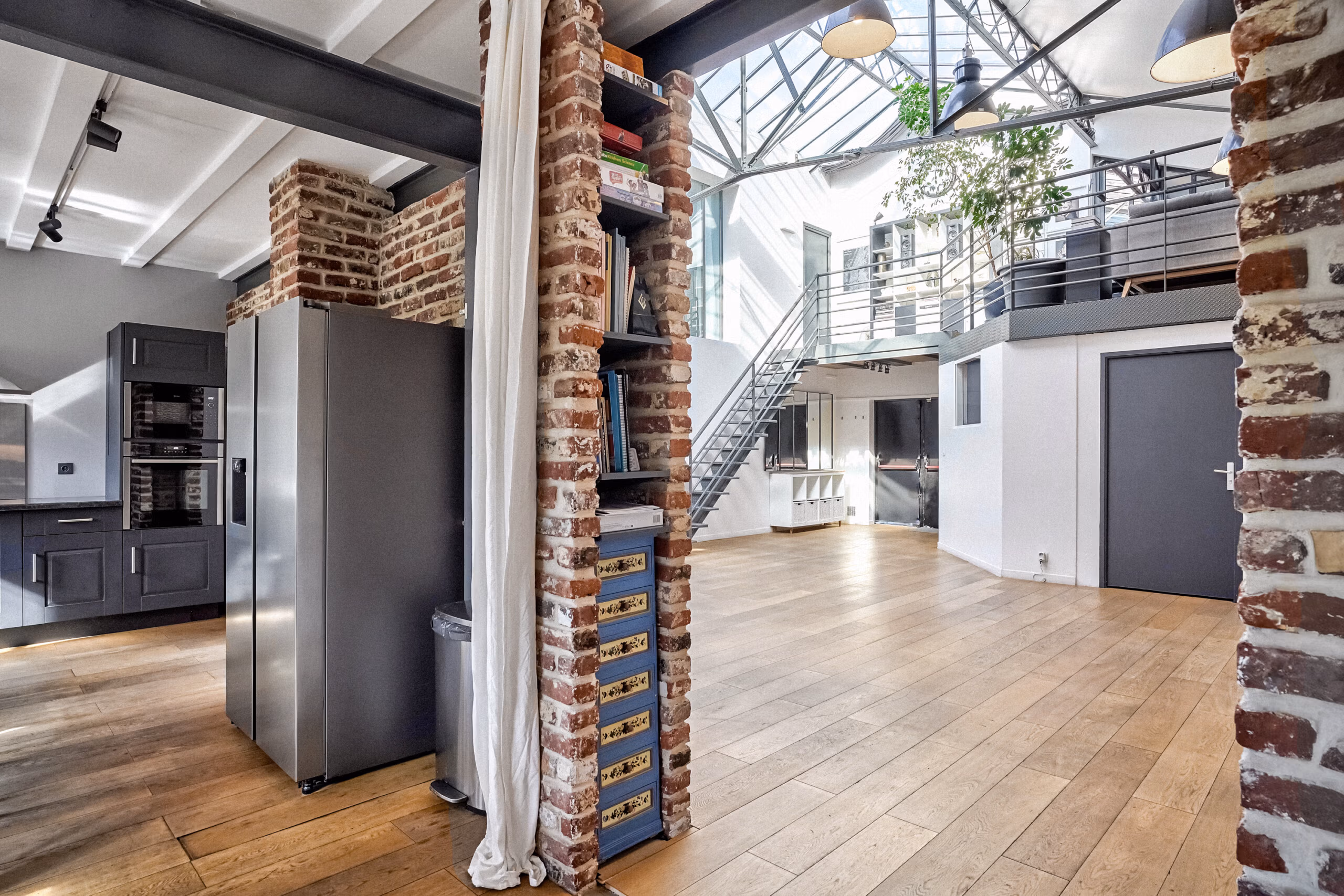 Vue traversante du duplex avec colonnes de briques apparentes, verrière cathédrale centrale, électroménager professionnel et parquet bois pour tournages décor industriel authentique