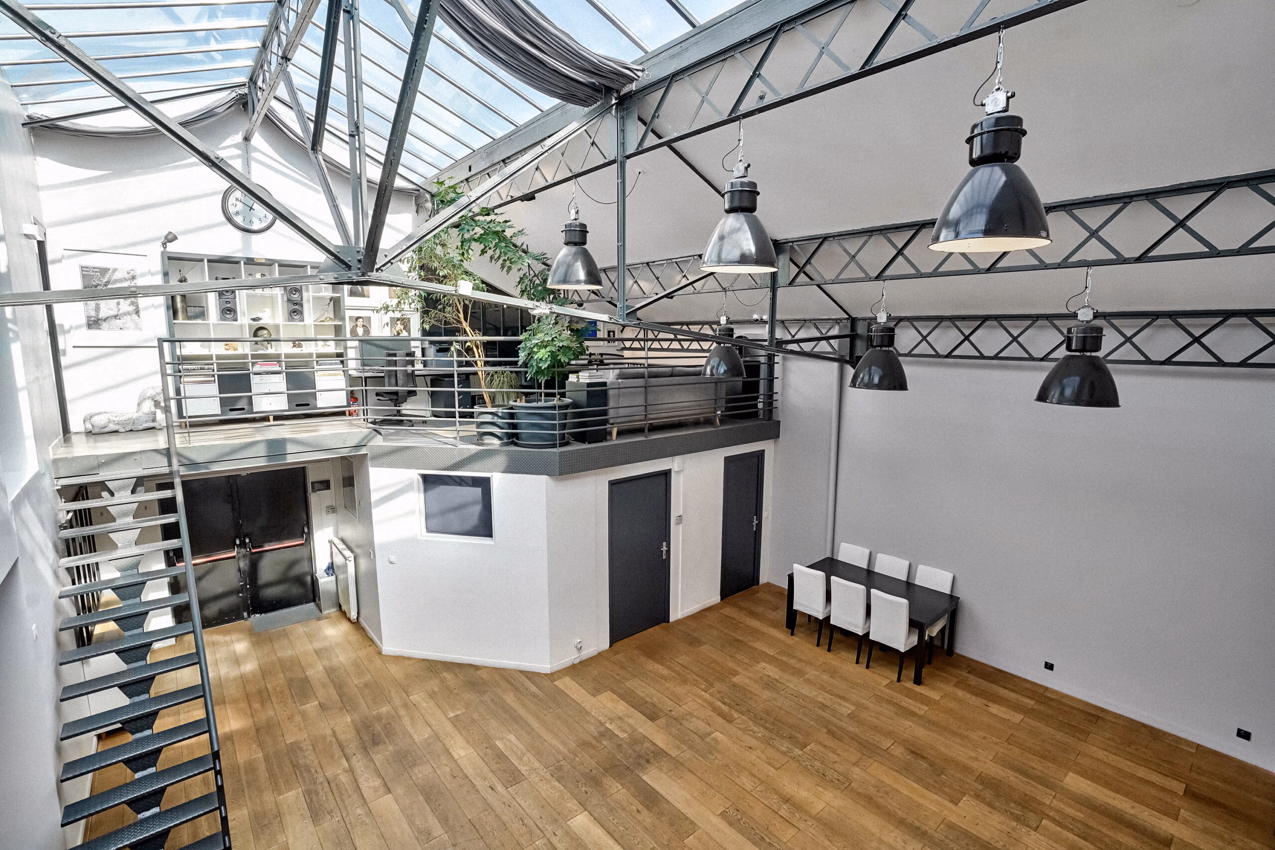 Vue panoramique du duplex avec table de réunion, verrière cathédrale, luminaires industriels suspendus et végétation