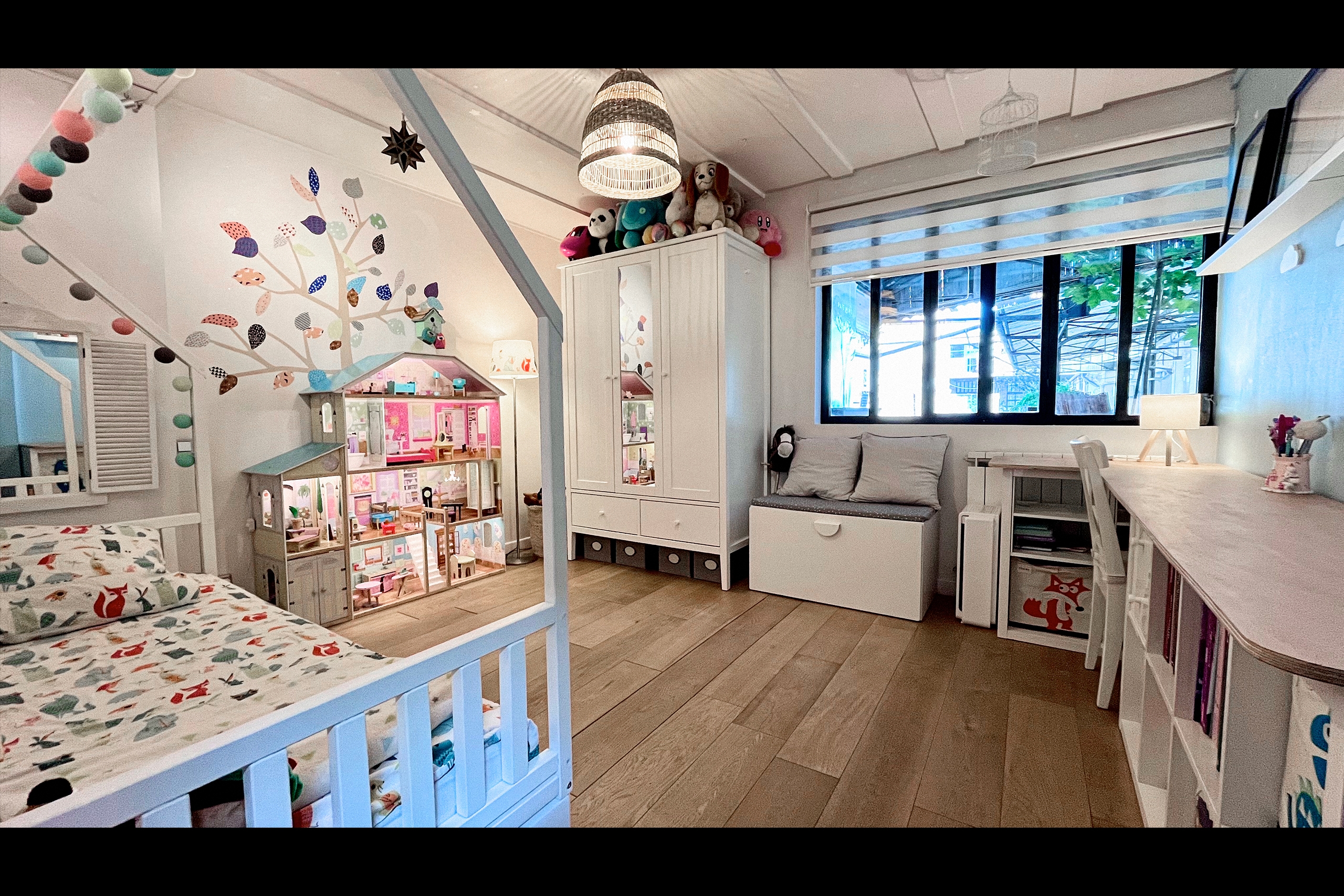 Chambre enfant avec lit cabane bois blanc, sticker mural arbre coloré, maison de poupée éclairée et suspension rotin tressée pour tournages enfance et shootings déco jeune public